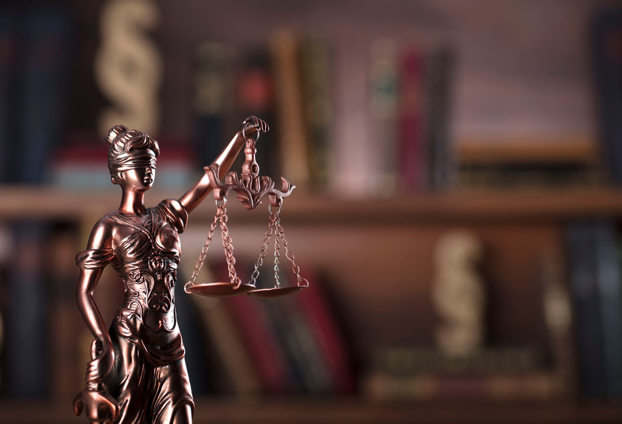 A bronze statue of Lady Justice holding scales, set against a blurred bookshelf filled with books.