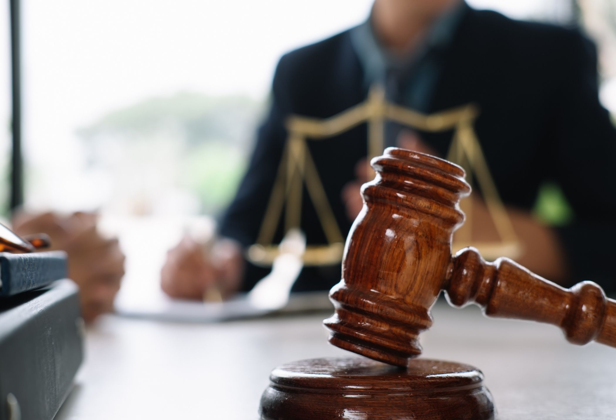 A wooden gavel rests on a table, with blurred figures of a lawyer and scales of justice in the background, hinting at a legal discussion.
