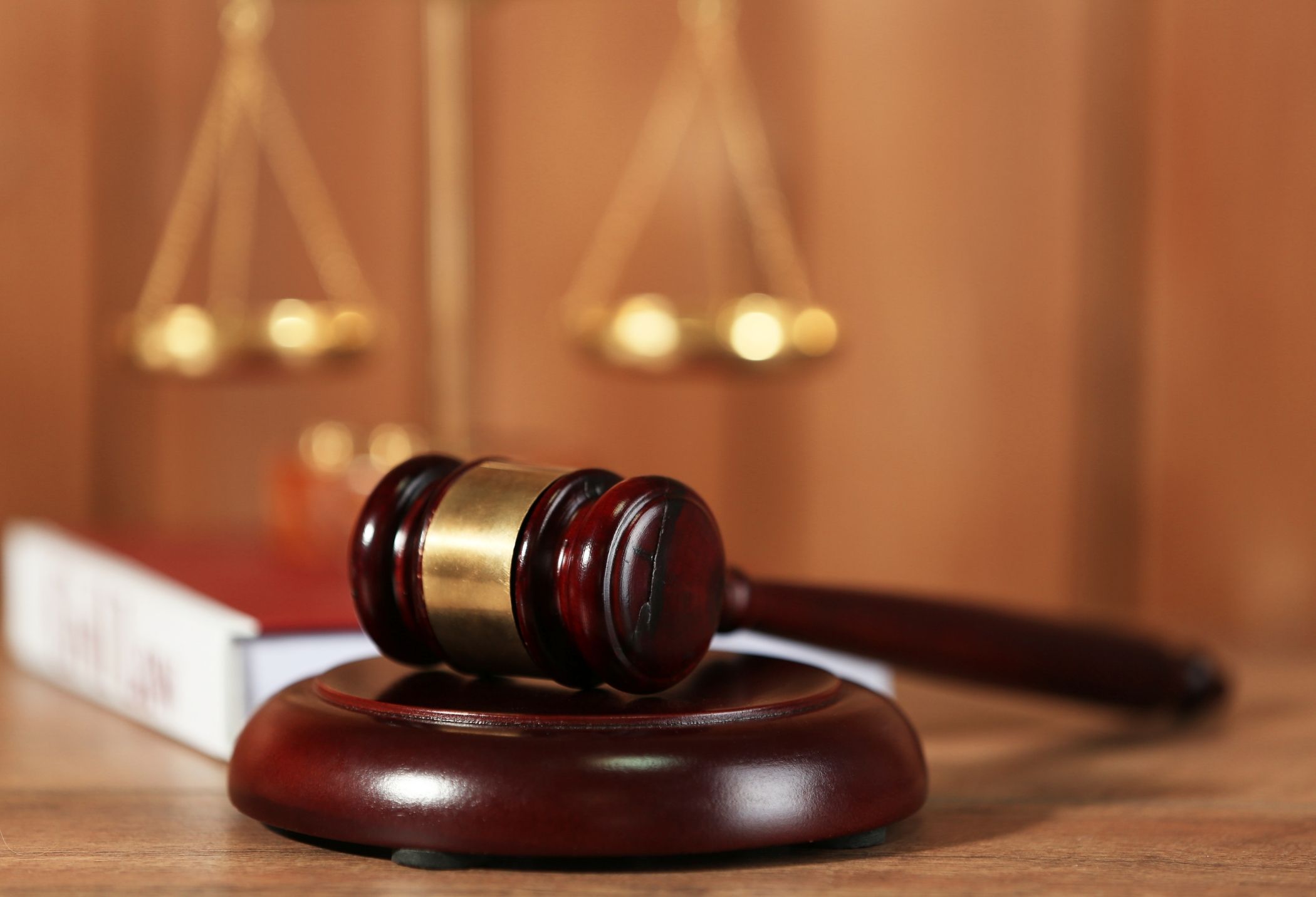 A wooden gavel rests on a round base, with a blurred legal scale and book in the background, symbolizing law and justice.