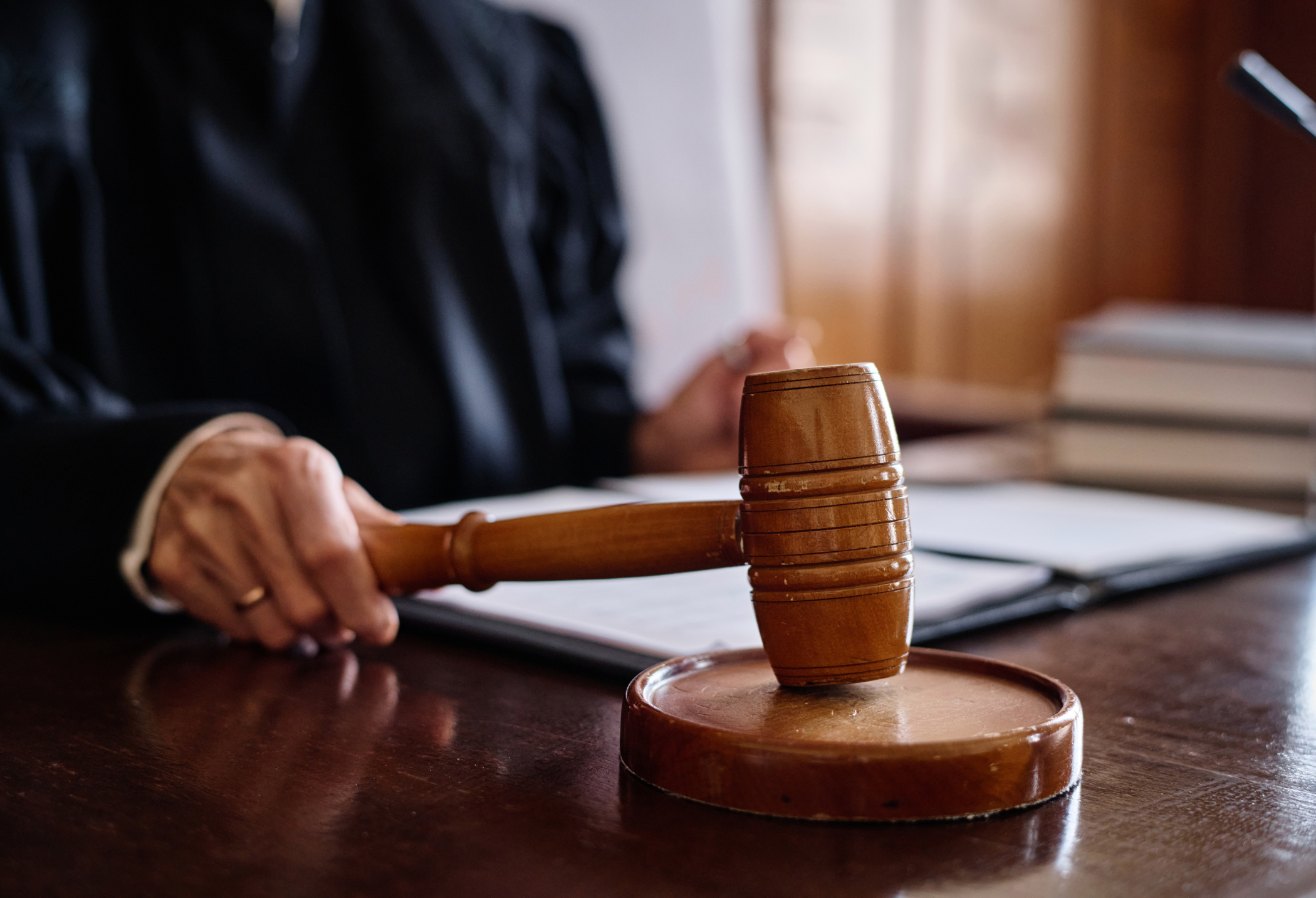 A judge's hand holds a wooden gavel over a desk, with legal documents and books in the background.
