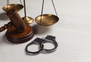 A wooden gavel and handcuffs beside a brass scale of justice on a light surface, symbolizing law and order.