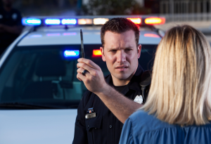 A police officer holds a pen while speaking with a person beside a police car, which has flashing lights.