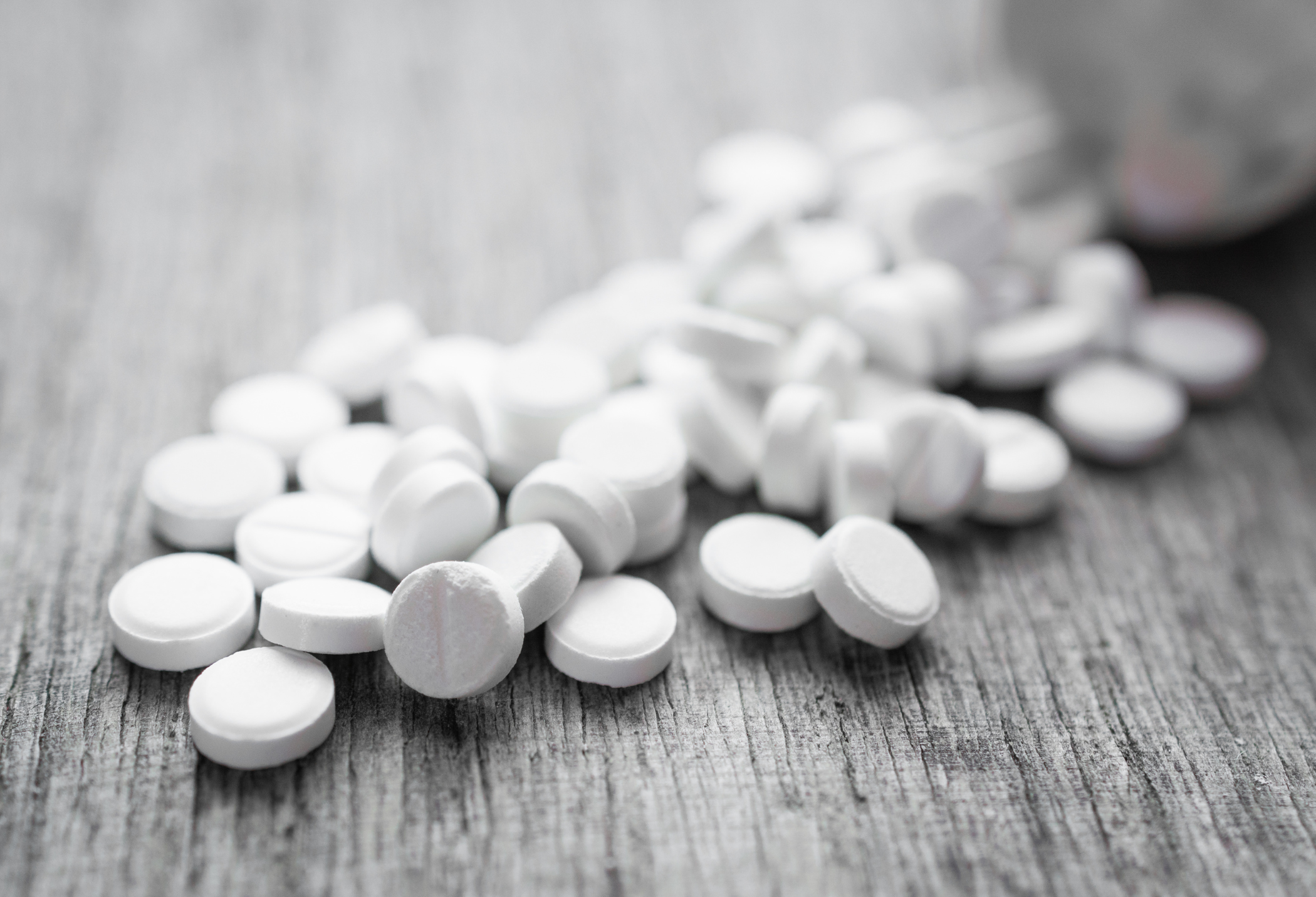 A close-up of scattered white tablets on a textured gray surface, some with engraved lines. A portion of a container is in the background.