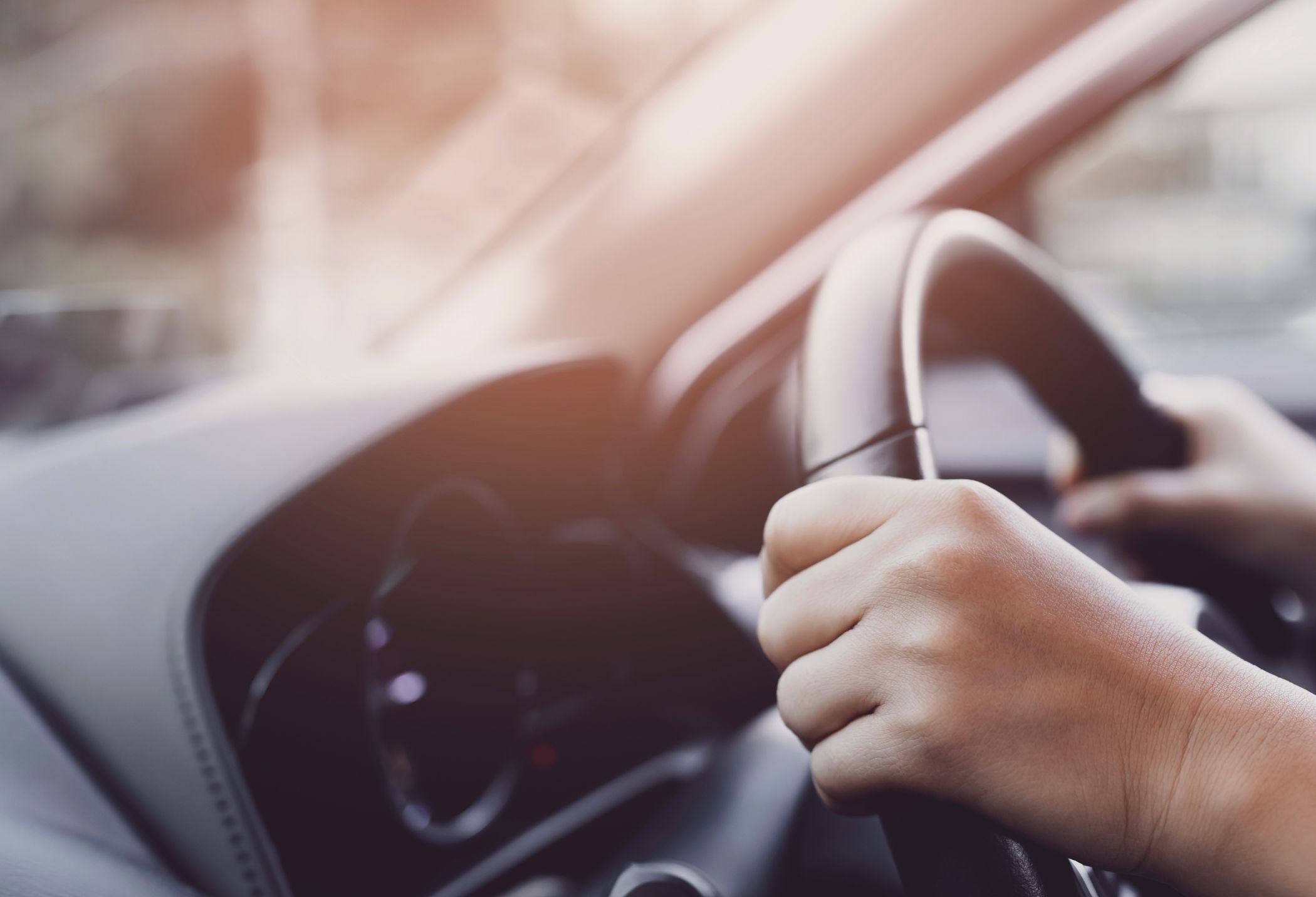Close-up of hands gripping a car steering wheel, with a blurred dashboard and soft light in the background.