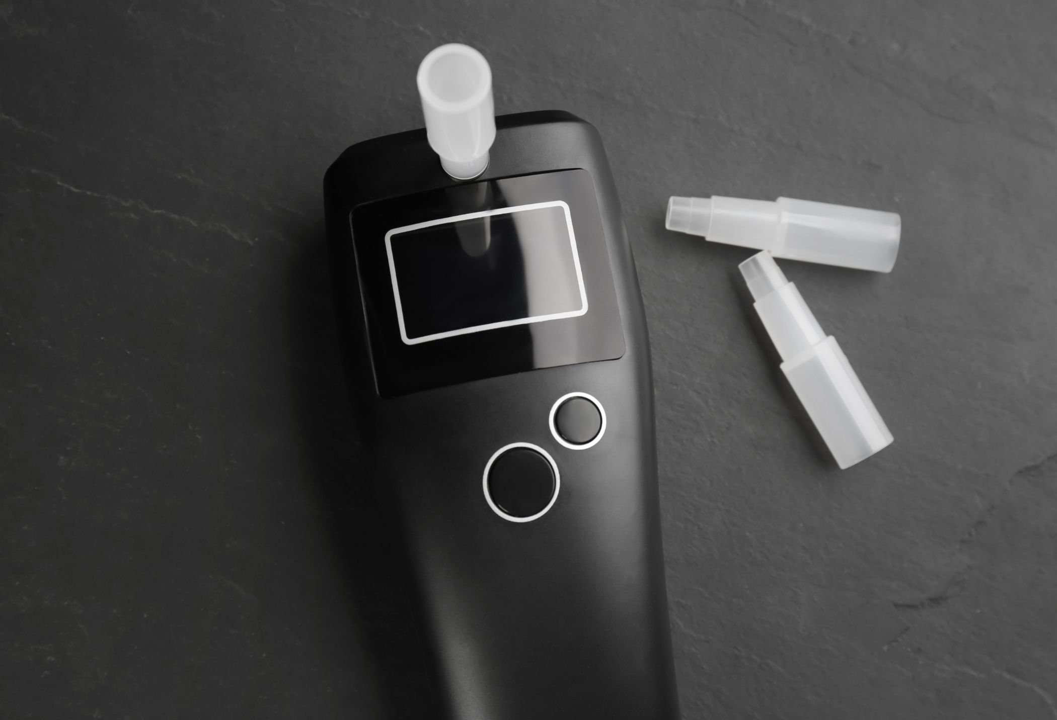 A black breathalyzer device with a digital display, featuring two buttons and three plastic mouthpieces nearby on a dark surface.