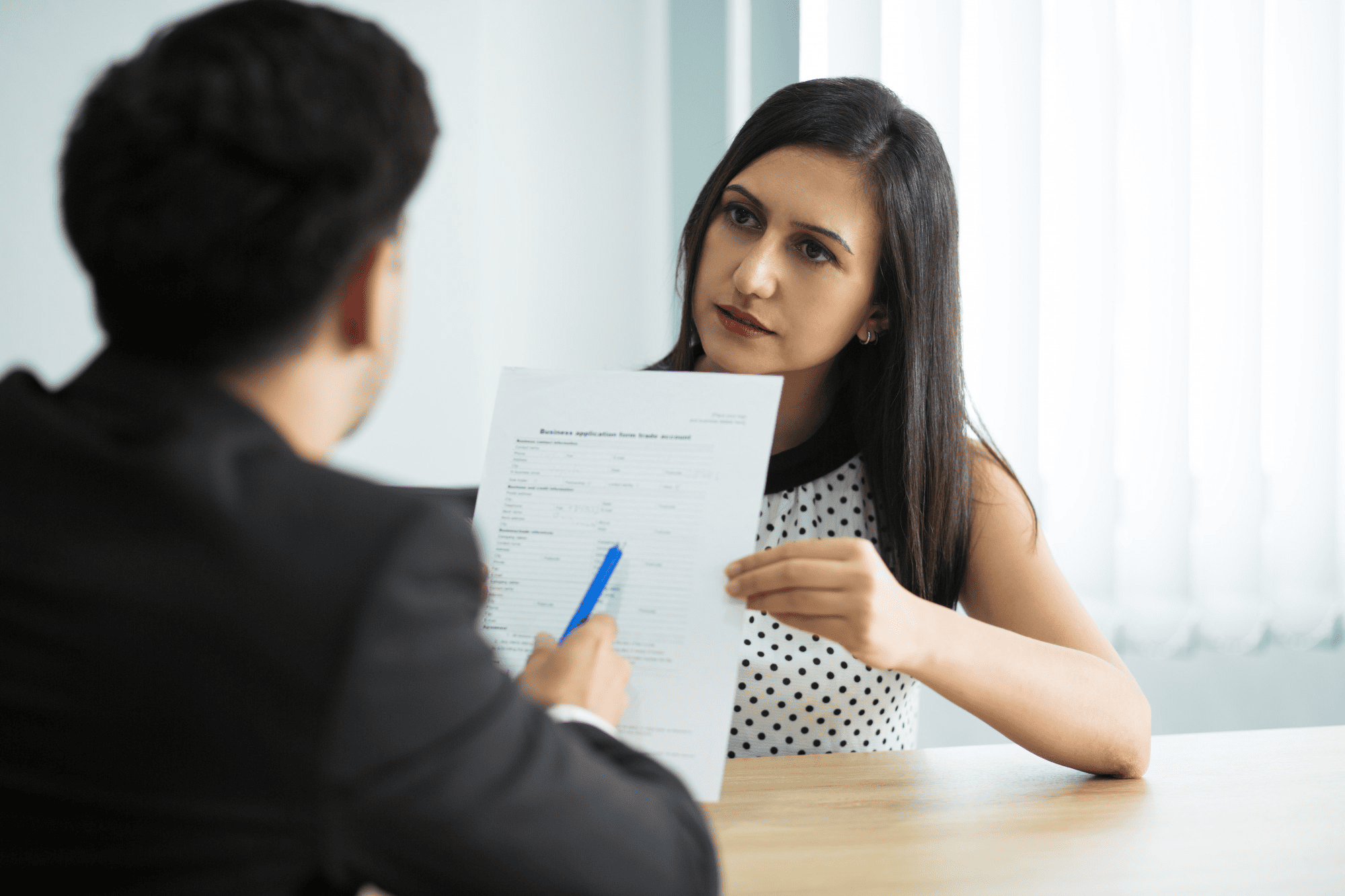 A woman in a polka dot top presents a business application during a meeting with a suited man at a table. Light streams in from behind.